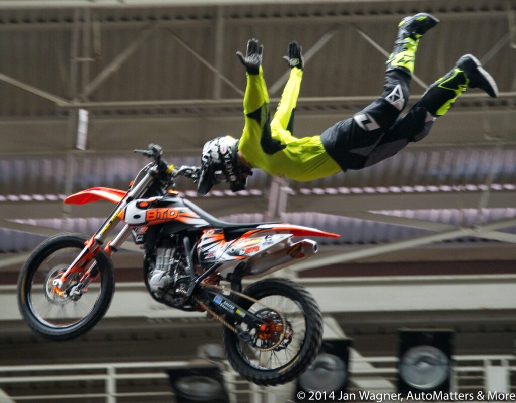 A man in yellow and black jumps over a motorcycle.