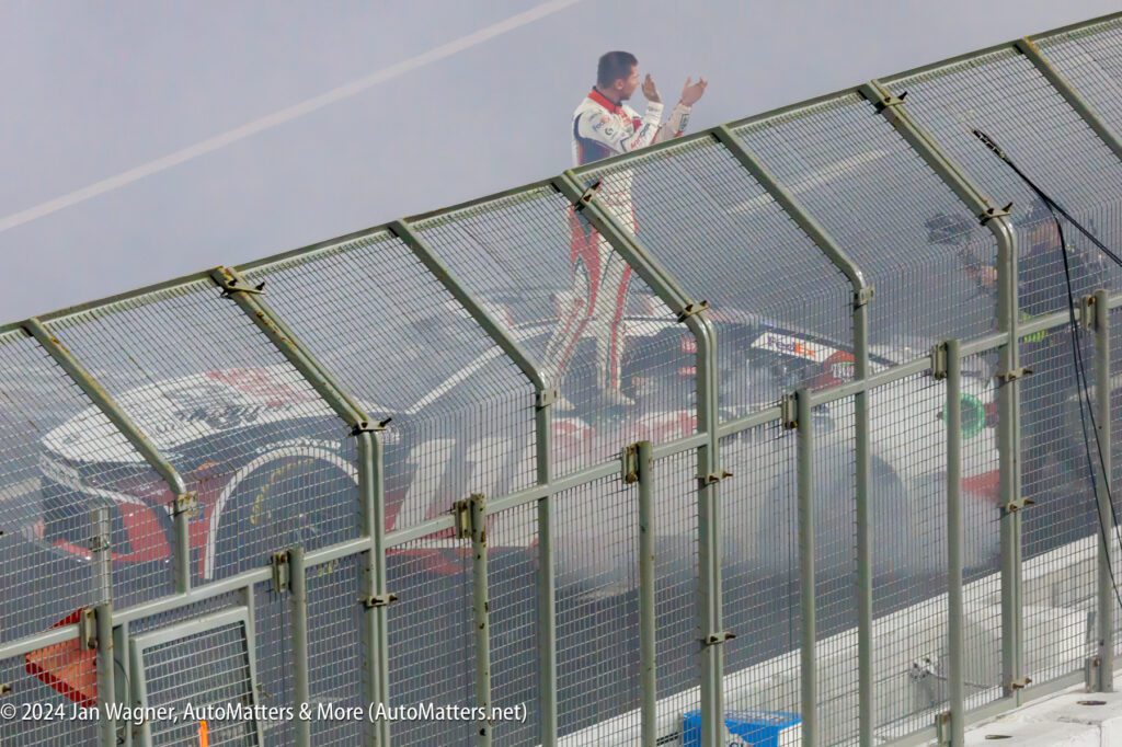 A nascar driver is standing on top of a fence with smoke coming out of his car.