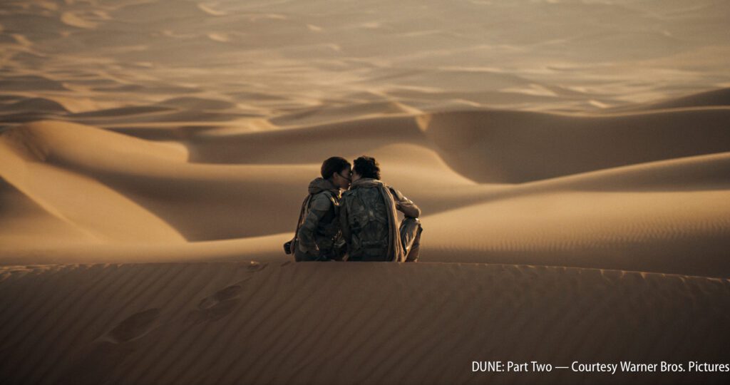 Two people sitting on top of a sand dune in the desert.
