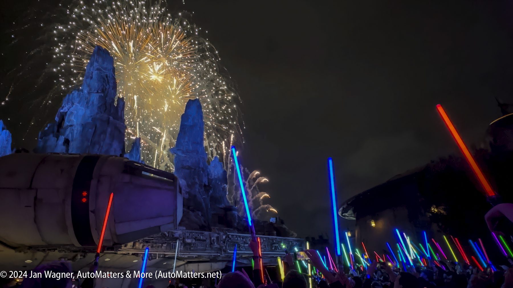 A crowd holding colored lightsabers looks up at a nighttime fireworks display set against rocky, futuristic structures.