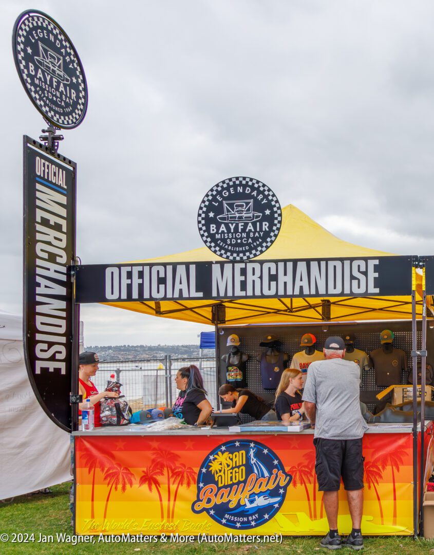 A man stands at an outdoor "Official Merchandise" booth for San Diego Bayfair, with staff and various hats and shirts on display.