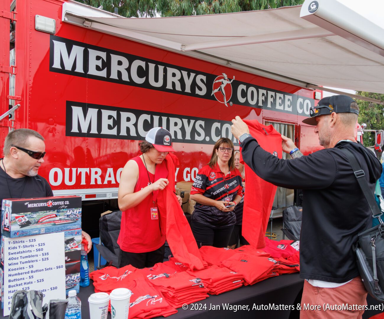 A group of people stand at a Mercurys Coffee Co. booth, handling red t-shirts near a price sign, with a red company truck in the background.