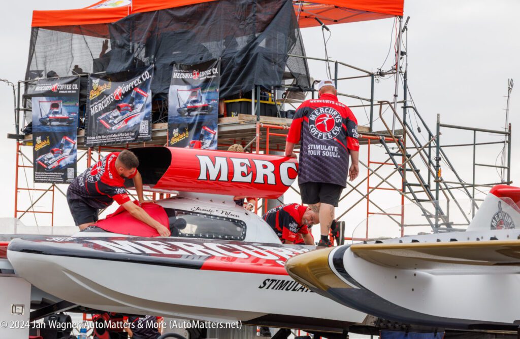 Three crew members work on a red and white hydroplane boat labeled “MERC” at a boat race event, with banners and equipment in the background.