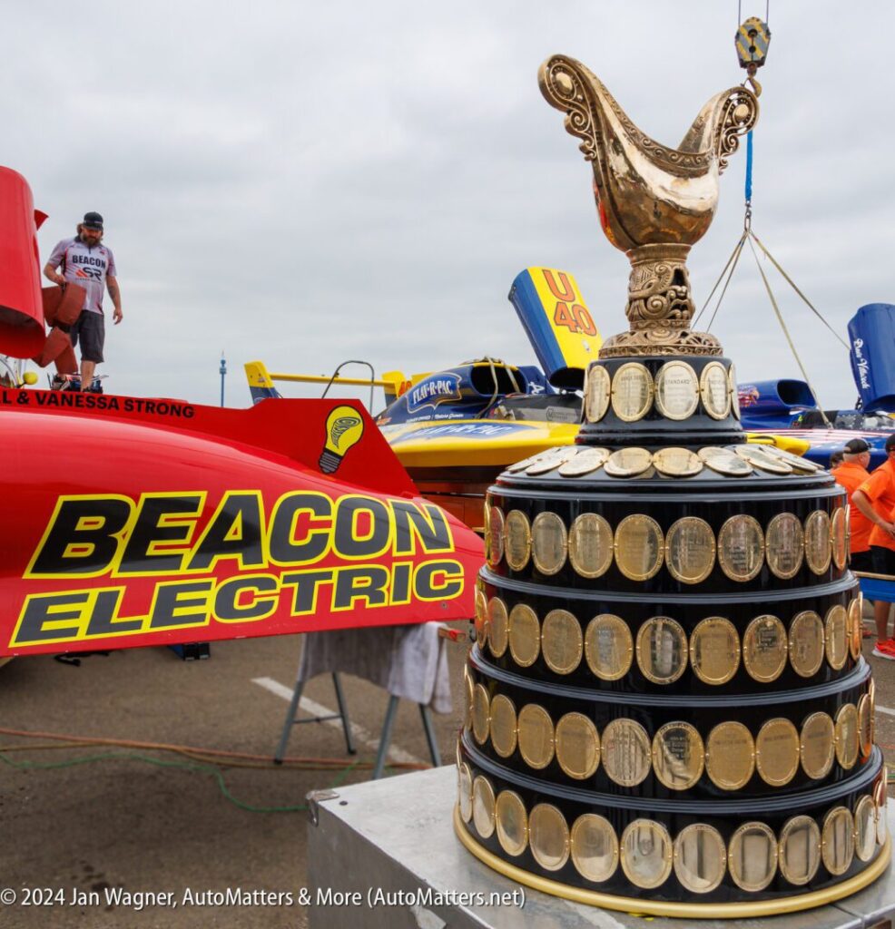 A large trophy with engraved plaques sits in front of racing boats and crew members at a dockside event.
