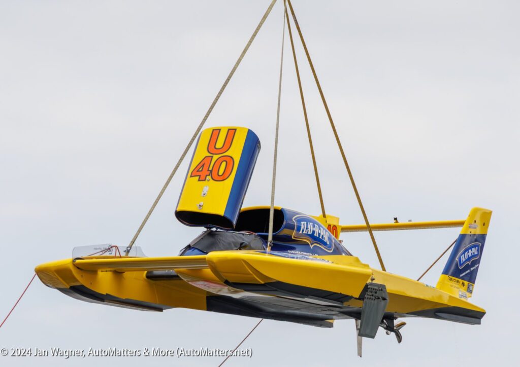 A yellow and blue hydroplane with "U-40" on the tail is being lifted by a crane, suspended in the air against a cloudy sky.