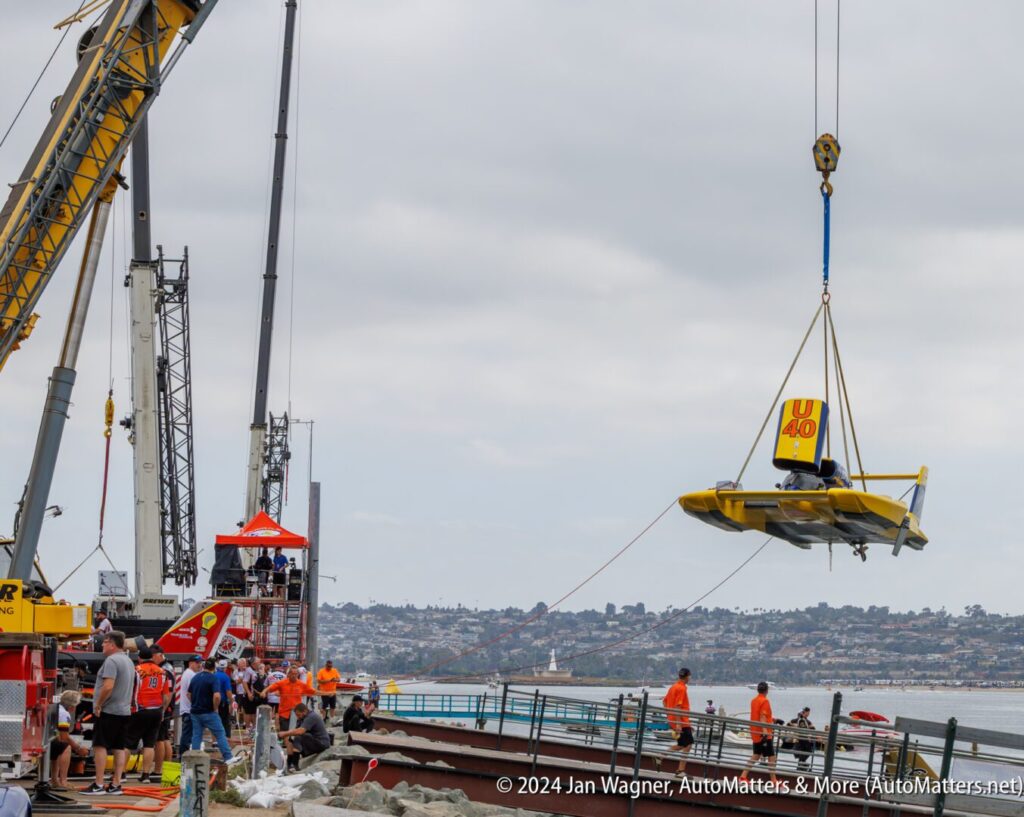 A crane lifts a yellow hydroplane boat labeled "U-40" over a dock area with workers and onlookers, with water and city in the background.