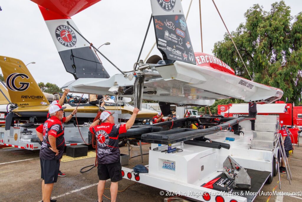 Team members in red shirts prepare a hydroplane boat with “Mercury Coffee Co.” branding, using a crane to lift it on a trailer at an outdoor event.