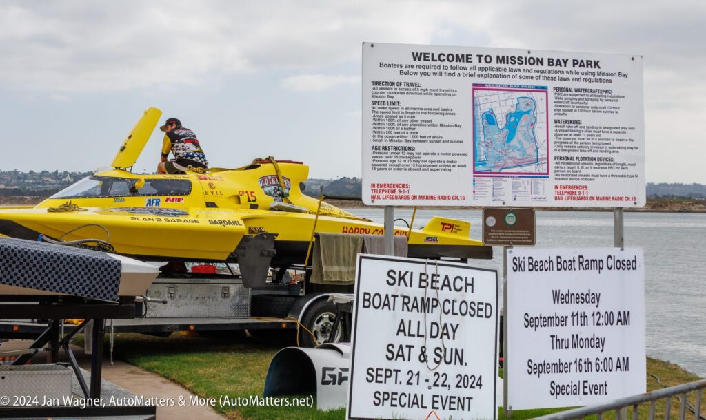 A yellow powerboat is being prepared near informational signs at Mission Bay Park, including notices about Ski Beach boat ramp closures. Water and cityscape are visible in the background.