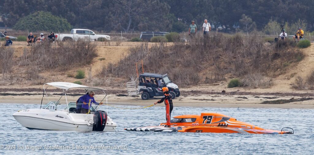 A person stands on an orange powerboat, holding a tow line connected to a white motorboat, with spectators and vehicles onshore in the background.