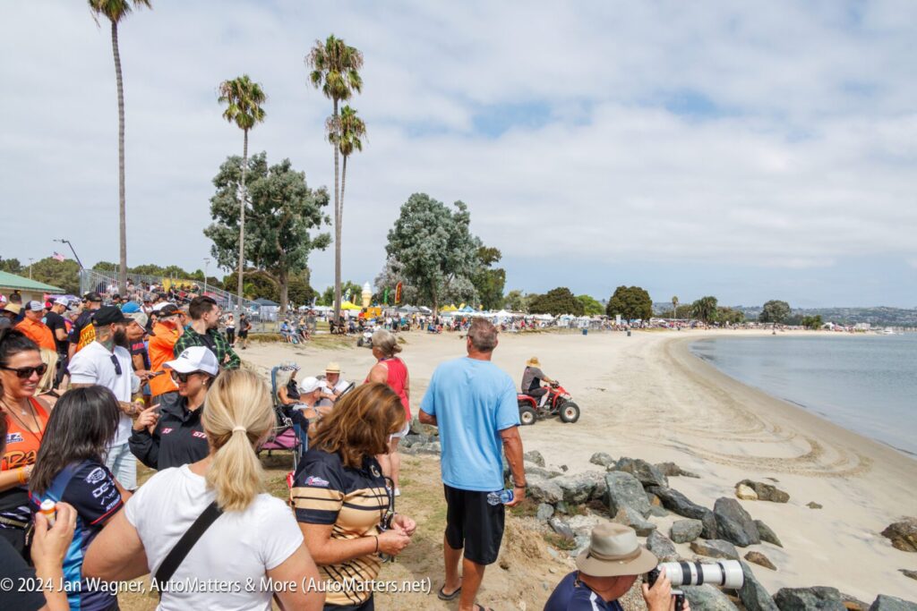A crowd gathers along a sandy shoreline near palm trees and rocks, watching an event. Some people take photos while a man drives an ATV along the beach.