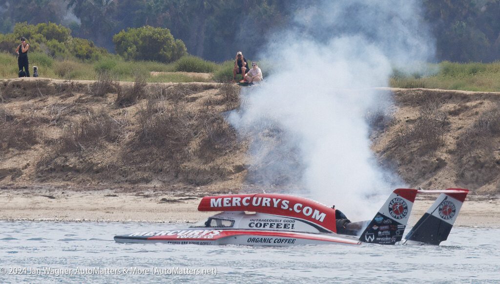 A hydroplane boat labeled "MERCURYS.COM" emits smoke on the water near a grassy shore, with two people sitting and watching in the background.
