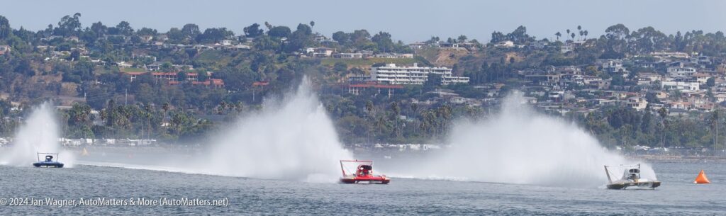 Three hydroplane boats race on a body of water, creating large sprays, with a hillside town visible in the background under a clear sky.