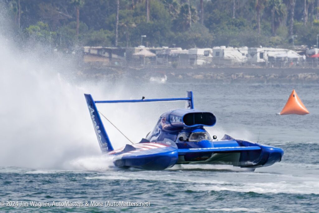 A blue hydroplane speeds across the water, creating a large spray, with an orange buoy and parked RVs visible in the background.