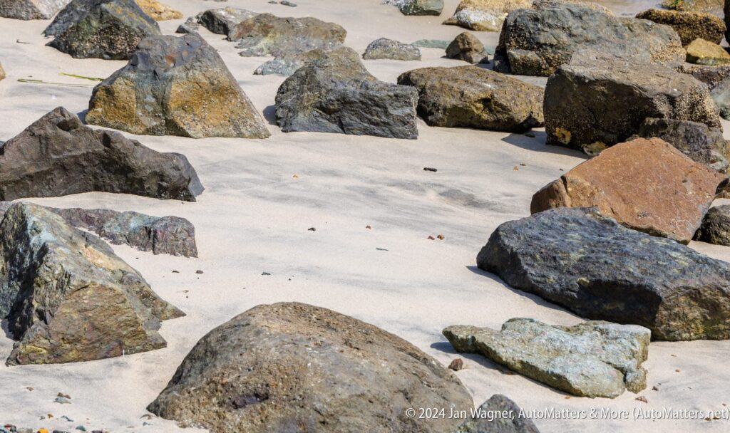 Large rocks scattered across a sandy beach with some small pebbles and the shadow of the rocks visible on the sand.
