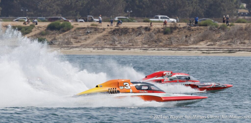 Three powerboats, numbered 3 and 93, race side by side at high speed, creating large sprays of water, with spectators and trees visible in the background.