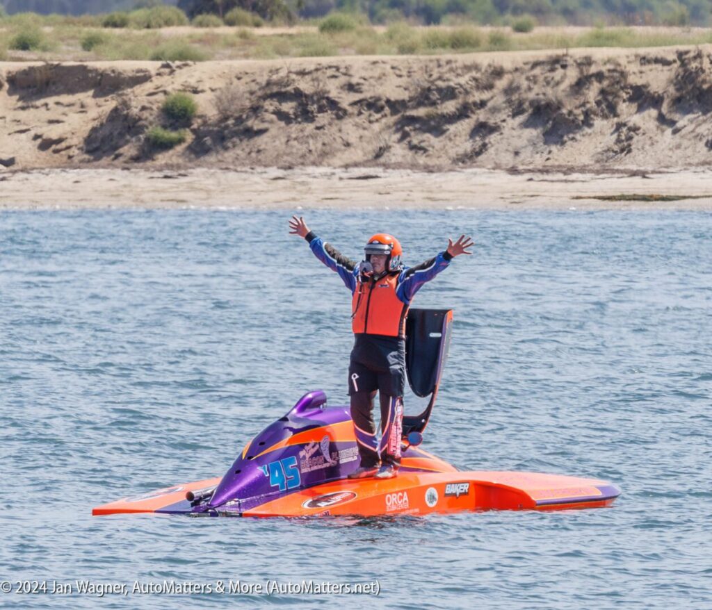 A person in racing gear and a life vest stands with arms raised on a purple and orange speedboat floating on water, near a sandy shoreline.