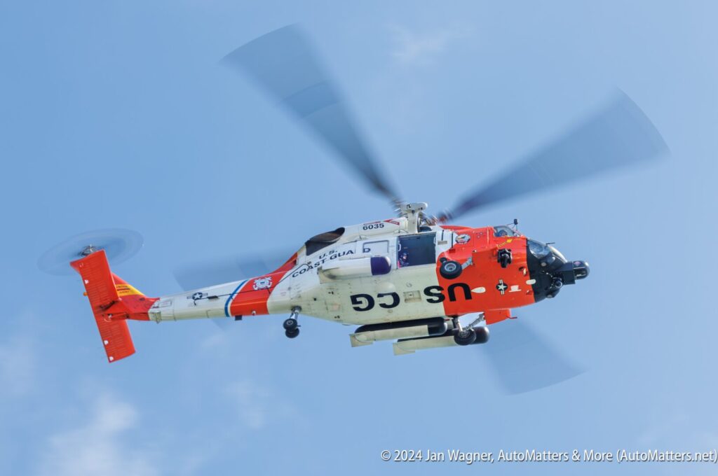 A U.S. Coast Guard helicopter in flight against a blue sky, with its rotors in motion and distinct red, white, and blue markings visible.