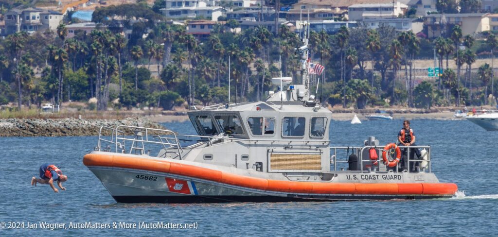 U.S. Coast Guard boat on the water with one person diving in and another standing on deck; shoreline with buildings and palm trees in background.