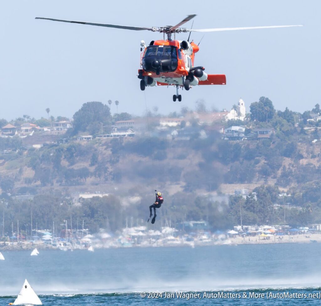 A helicopter hovers over water as a person in a wetsuit is being lowered by a cable during a rescue demonstration near the shore.