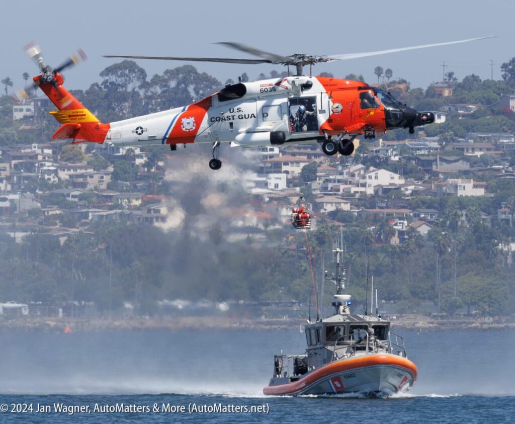 A U.S. Coast Guard helicopter performs a rescue drill over water, lowering a crew member toward a Coast Guard boat near a shoreline with houses in the background.