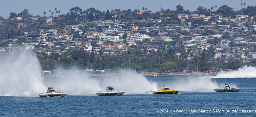 Four speedboats race on a body of water, creating large sprays behind them, with a hillside neighborhood visible in the background.