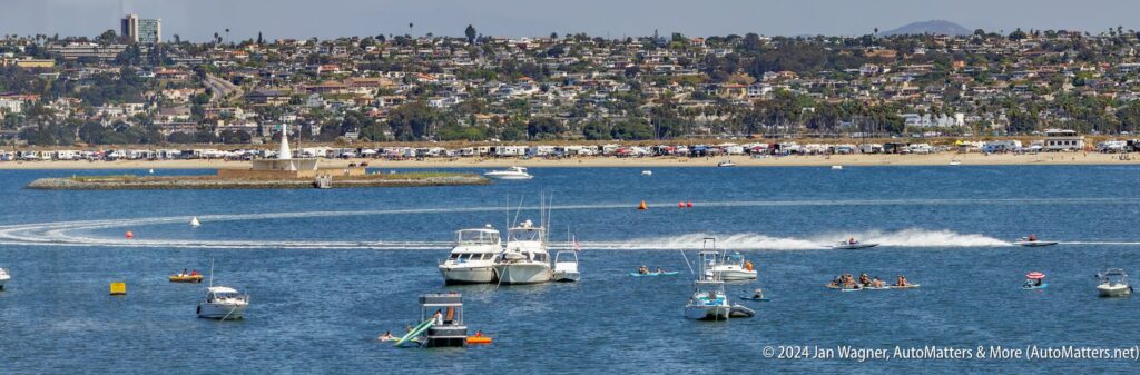 Boats, personal watercraft, and kayaks are active on a bay with a cityscape and parked vehicles visible along the distant shoreline.