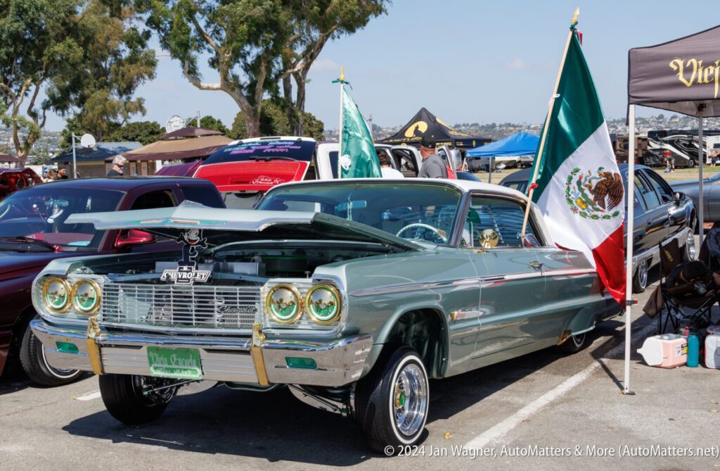 A vintage green Chevrolet Impala lowrider with a raised front end, displayed at a car show next to a large Mexican flag.