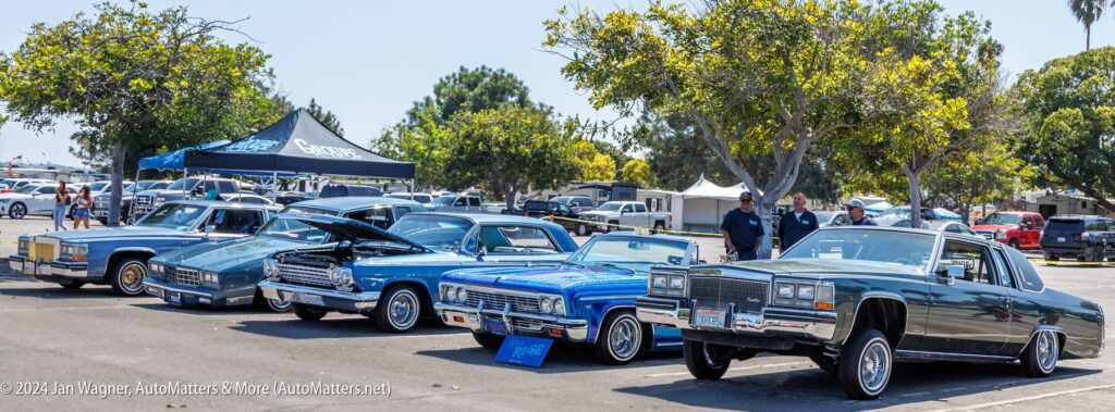 Four classic lowrider cars with raised suspensions are parked in a row at an outdoor car show, with trees, tents, and people in the background.