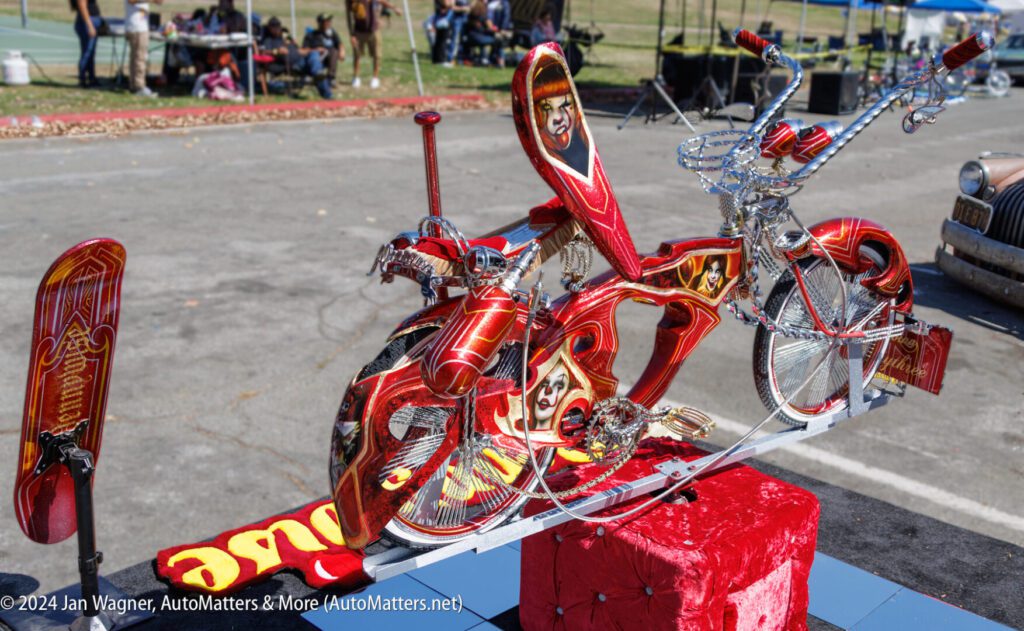 A custom red lowrider bicycle with intricate chrome details and airbrushed portraits is displayed on a raised stand at an outdoor event.