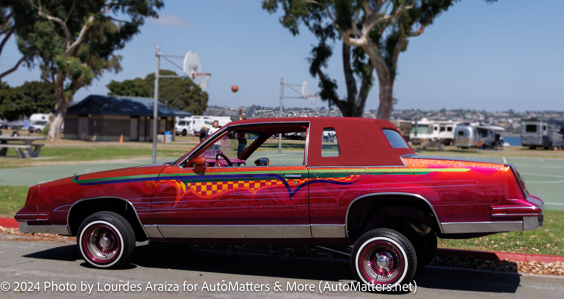 A custom lowrider car with vibrant red and pink paint, raised front wheels, and detailed designs, parked near a basketball court and trees by the water.