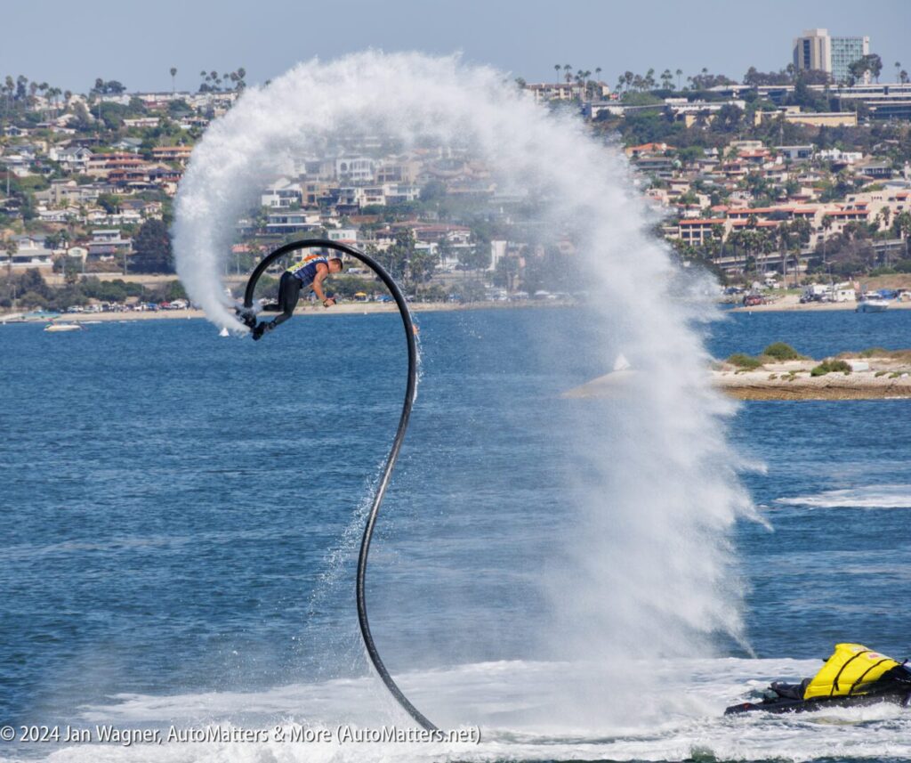 A person uses a water-powered jetpack to perform a high arc above the ocean, creating a large spray, with a coastal city in the background.