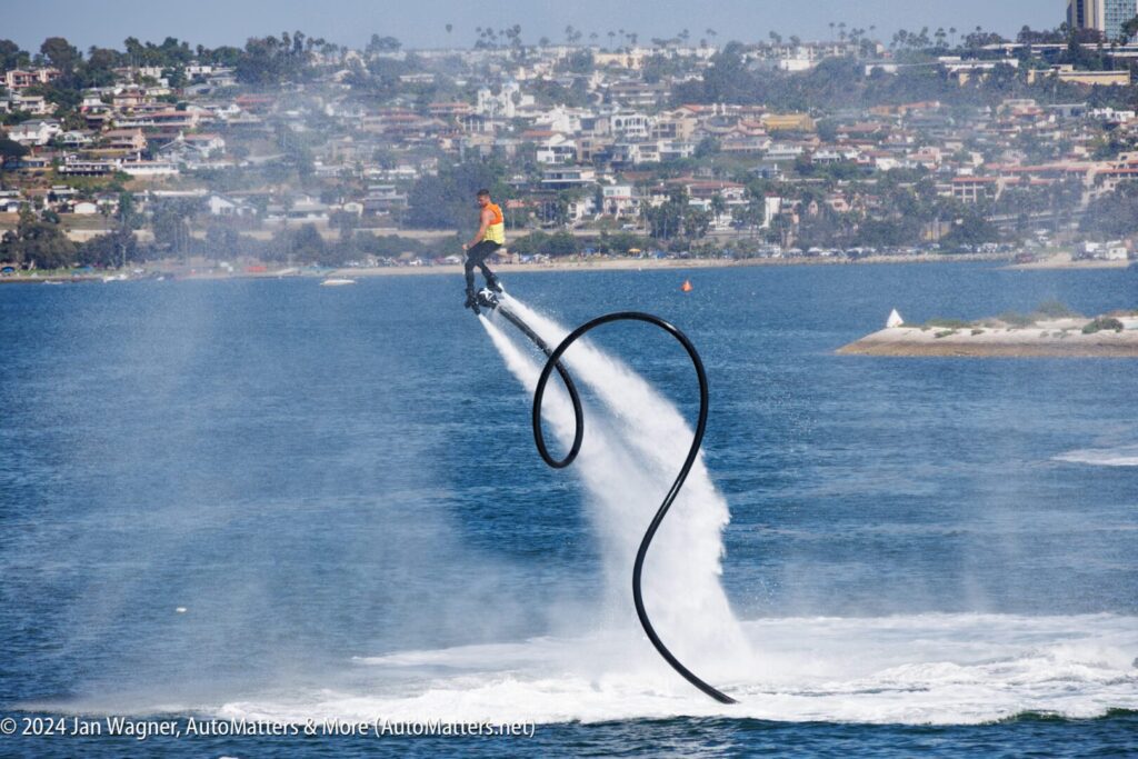 A person uses a water-powered jetpack, creating an arc of water above a bay, with a cityscape visible in the background.