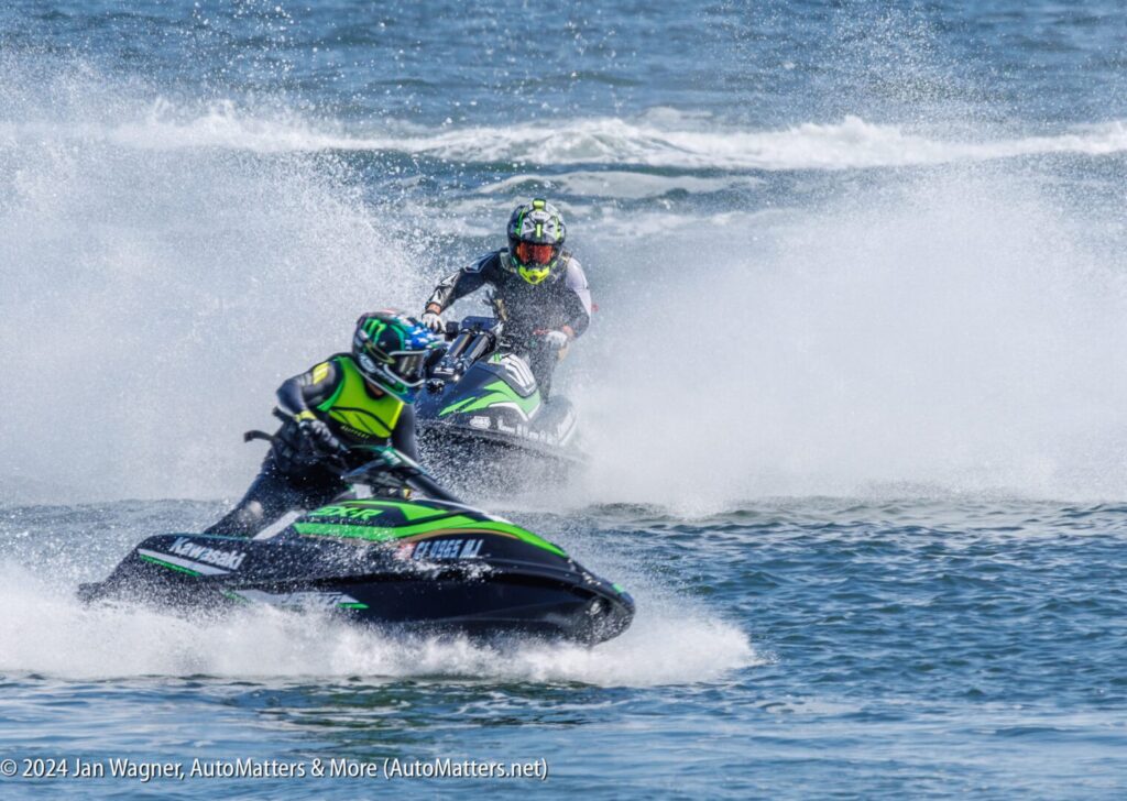 Two people ride Kawasaki jet skis at high speed on open water, creating spray as they maneuver during a race or competition.
