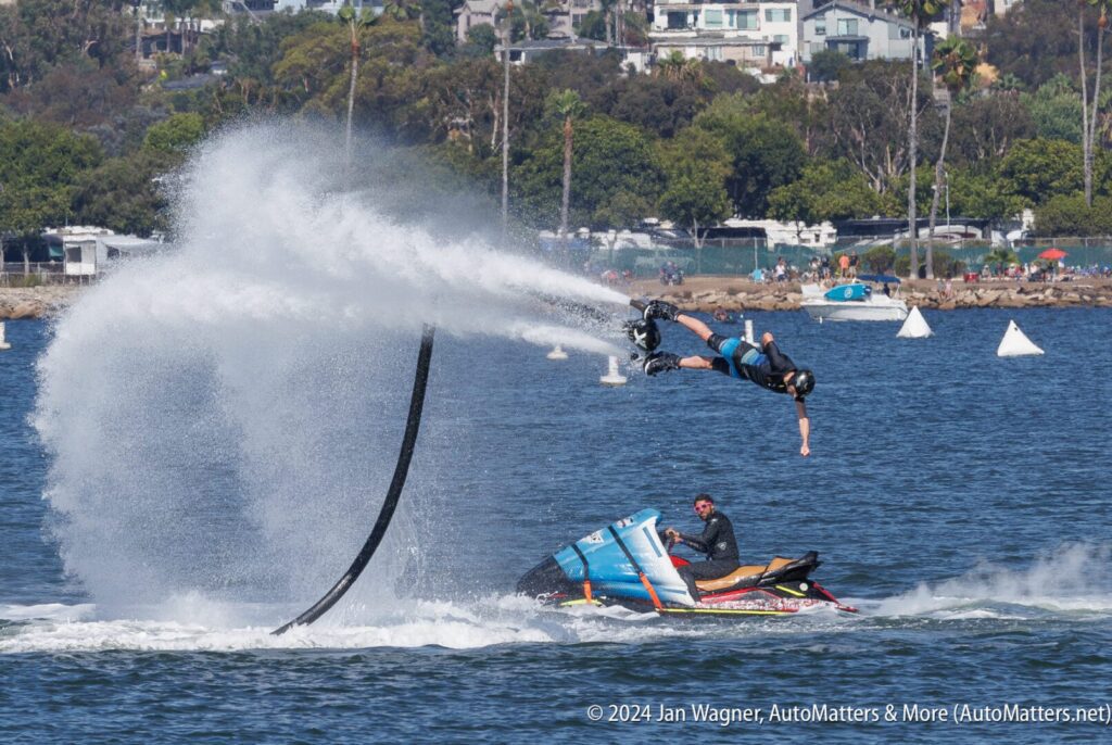 A person operating a water-powered jetpack hovers above the water, with another person falling off a jet ski below, both surrounded by water spray.