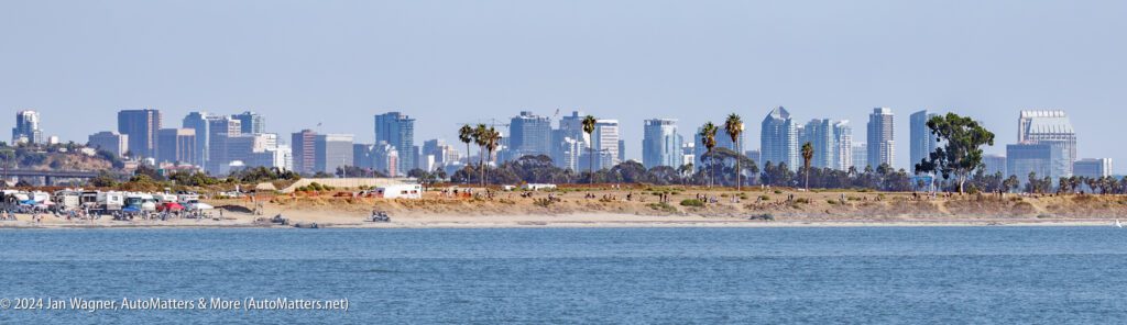 Panoramic view of a sandy shoreline with scattered trees and RVs, with a city skyline and tall buildings in the background across the water.