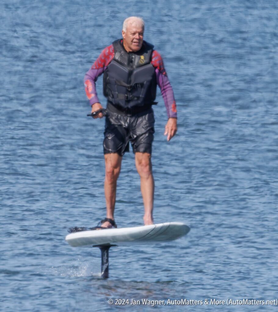 An older man wearing a life vest rides an electric hydrofoil surfboard above the water on a sunny day.