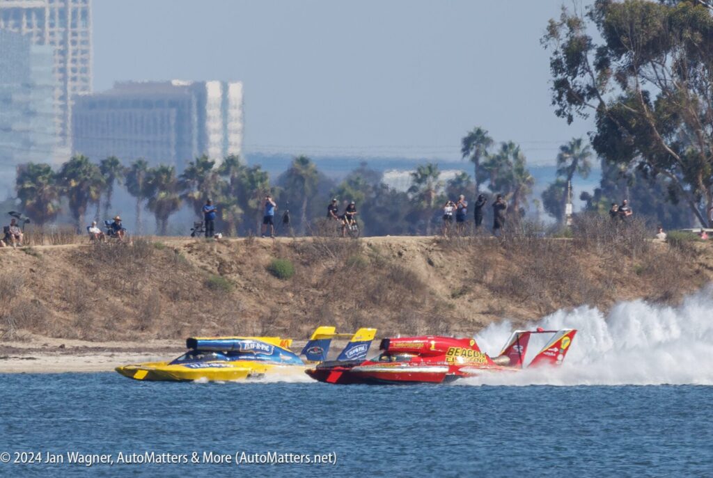 Three colorful hydroplane boats race side by side on the water near a shoreline with spectators and trees in the background.