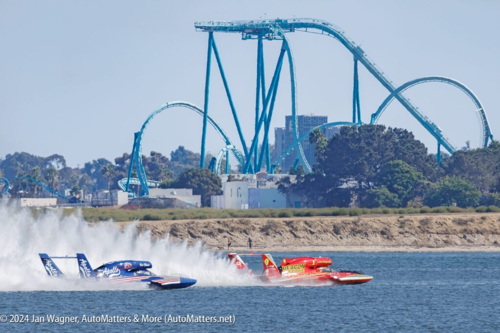 Three hydroplane race boats speed across a body of water, with a roller coaster visible in the background.