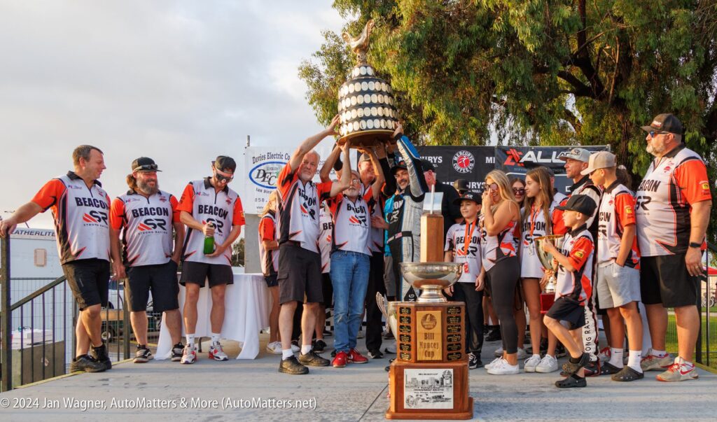 A sports team in matching uniforms celebrates on stage as two members lift a large trophy; other trophies and people are visible nearby.