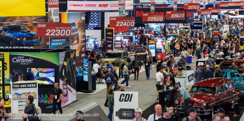 Crowded convention hall with booths, cars on display, and attendees walking and observing exhibits.