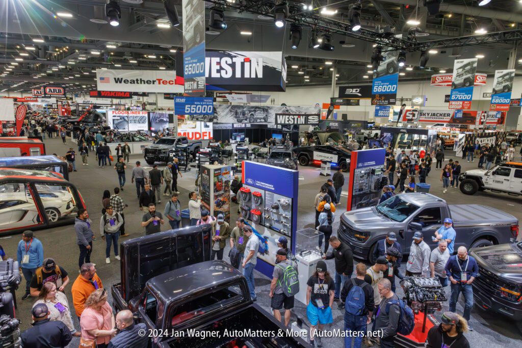 A bustling automotive trade show with attendees exploring various car exhibits and displays in a large exhibition hall.