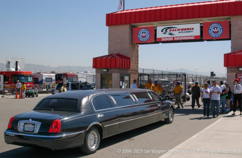 A black stretch limousine is parked at the entrance of California Speedway, where people are gathered near the gate under a sign that reads “GOOD MORNING.”.