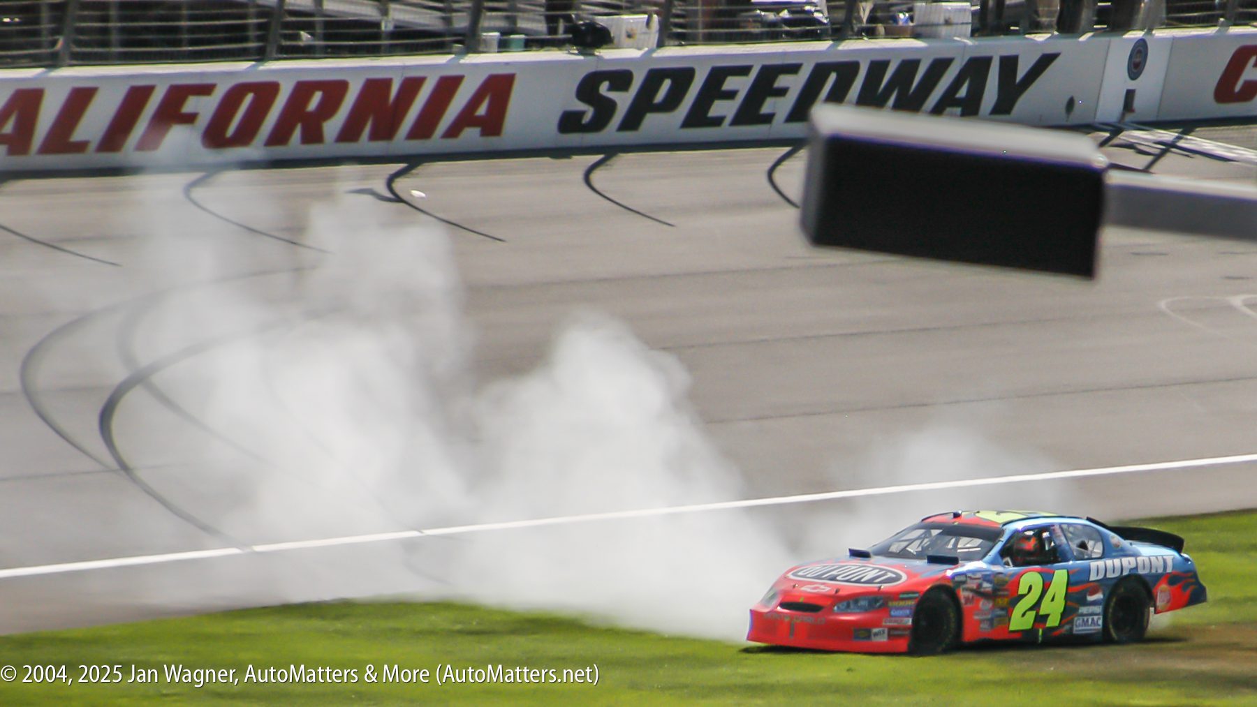 A race car with the number 24 leaves a trail of smoke as it spins off the track onto a grassy area at California Speedway.