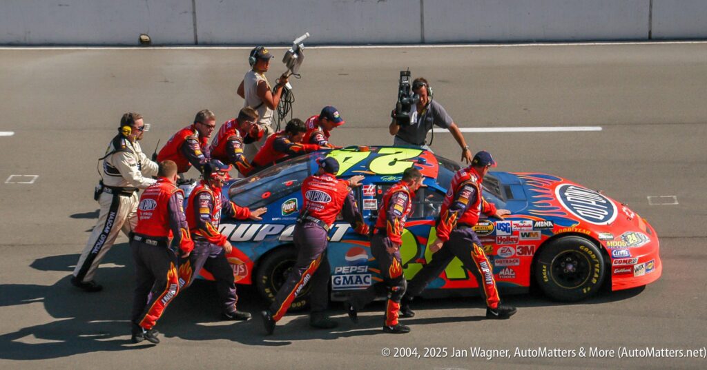 A NASCAR pit crew in red uniforms services a blue and red #24 car on pit road while a camera crew films the action.