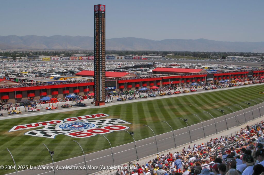 View of a racetrack with "Auto Club 500" painted on the infield, a scoreboard tower, pit garages, race cars, and a large crowd in the grandstands.