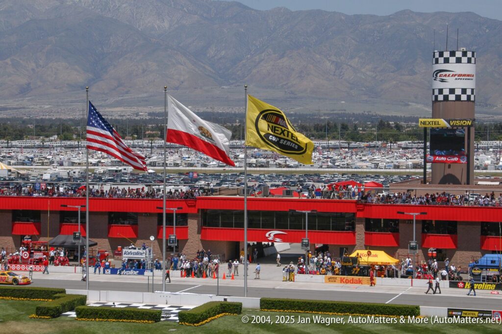 Three flags—American, California state, and a yellow Nextel flag—fly in front of a racetrack building with mountains and a large parking lot in the background.