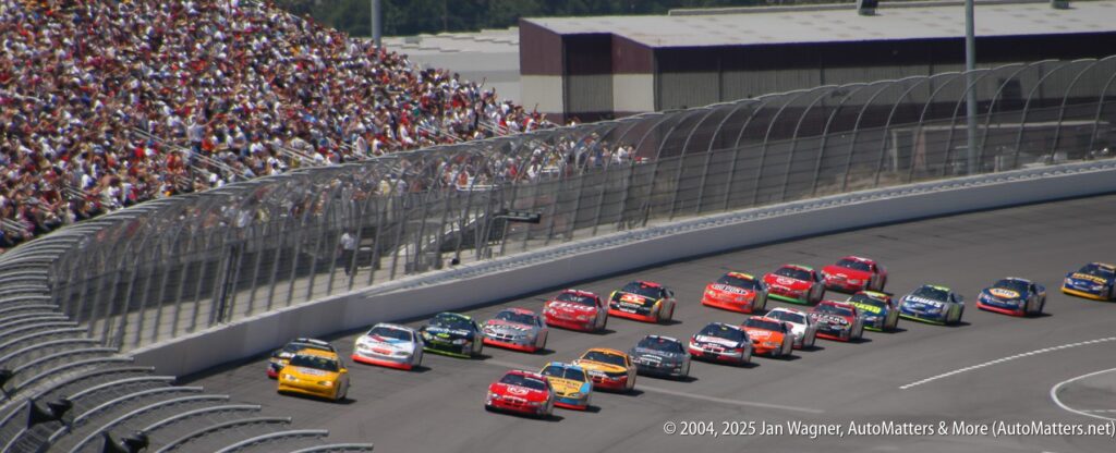 A group of stock cars races around a turn on a paved track as a large crowd watches from the grandstands.
