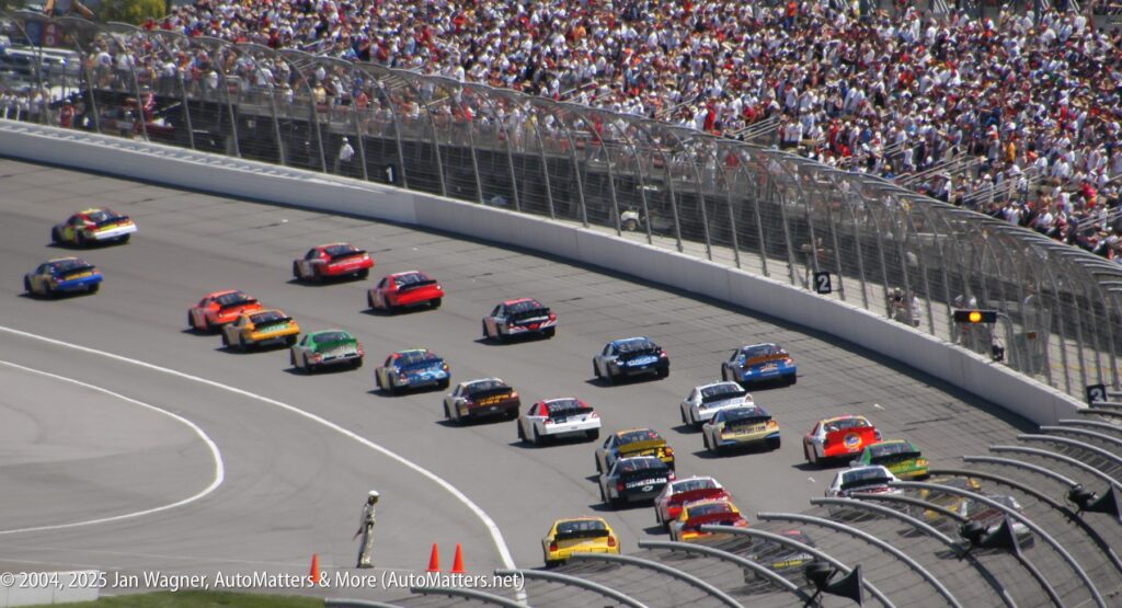 Stock cars race closely together on a banked track, with a packed grandstand of spectators in the background under a sunny sky.