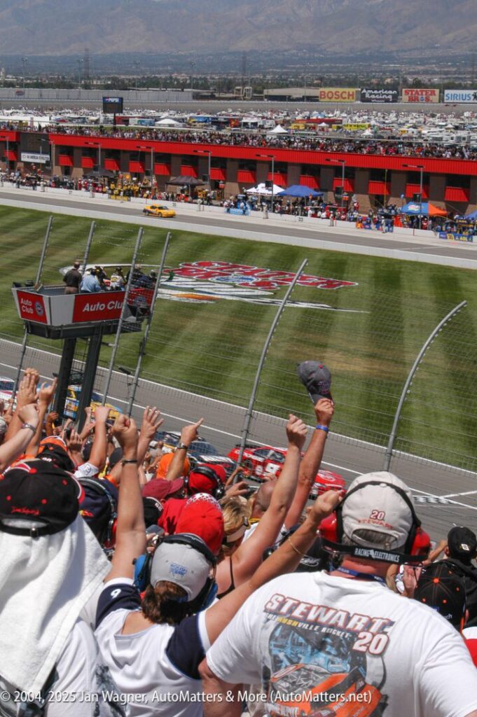 Crowd of fans in the grandstands cheer as race cars speed past the finish line at the Auto Club Speedway during a NASCAR event.