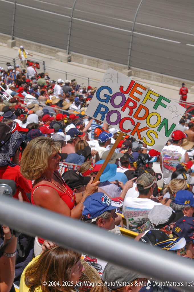A woman in a stadium crowd holds a colorful sign that reads "JEFF GORDON ROCKS" during a car racing event.